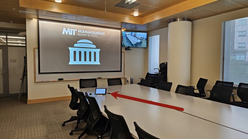 Picture of a large conference room in building E62 pointing to the Zoom Room Controller touchpanel on the table